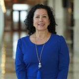 Melanie Shekita standing and smiling in a hallway with a blue dress on